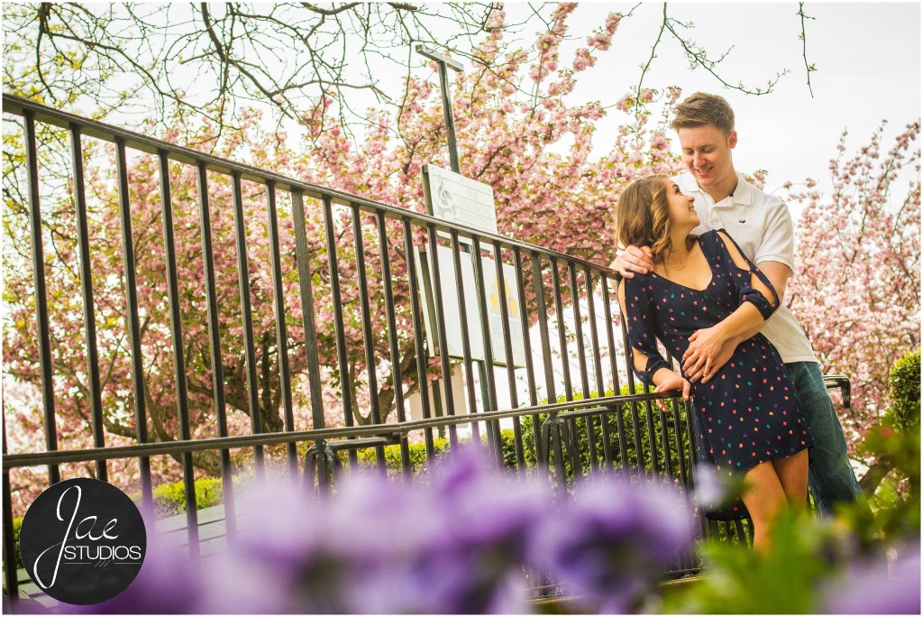 Hannah & Nick-41, Lynchburg Couple Session, Cherry Blossom, Black Metal Fence, Purple Flowers, Holding from Behind, Holding Hands, Looking at each other, Blue Patterned Dress, White Shirt, Brunette