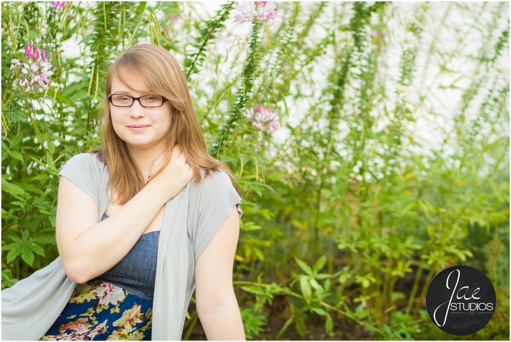 Lynchburg Senior Girl Emily 2014. Emily is relaxing on the ground with a blue flowered dress and a gray cardigan in front of pink flowers while perched on one arm. The other arm is resting on her opposite shoulder.
