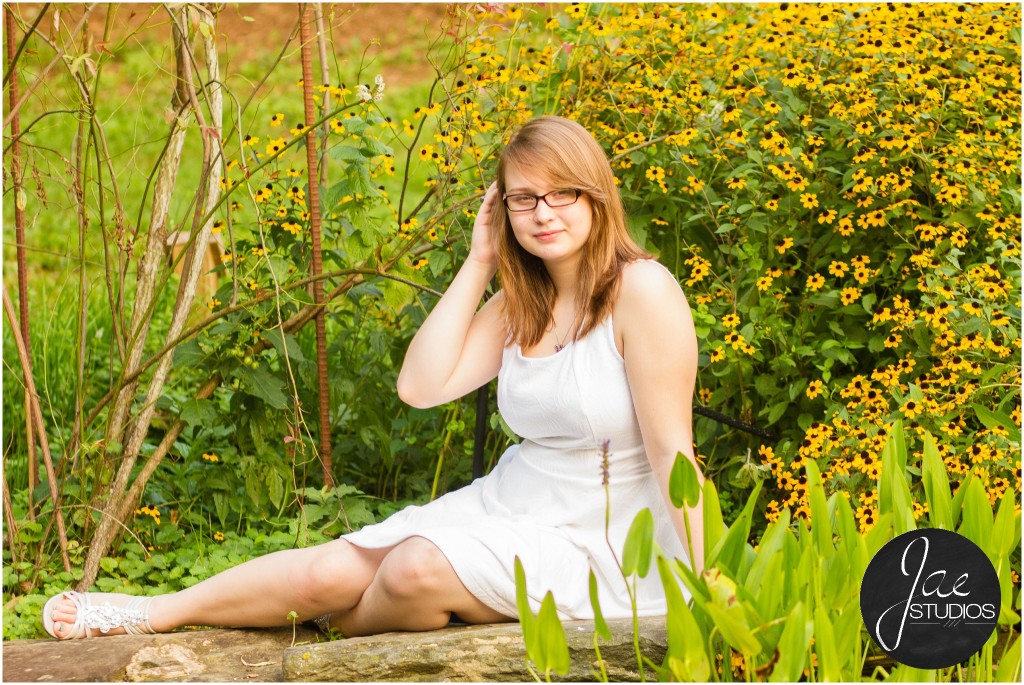 Lynchburg Senior Girl Emily 2014. Emily is sitting on a log in a white dress and white flats while surrounded by plants and yellow flowers.