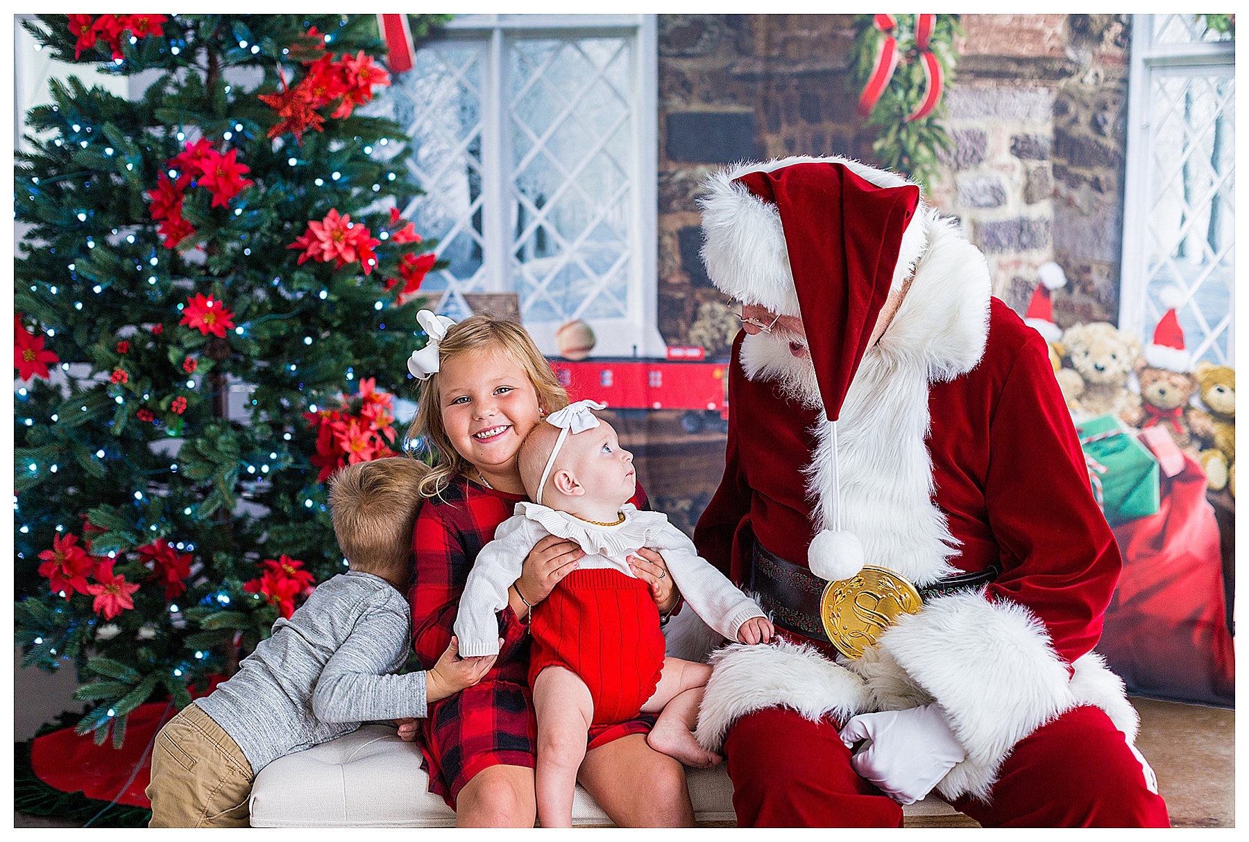A little girl smiling in front of a Christmas tree, while her little brother hides behind her and the little sister in her lap stares up mesmerized by Santa.