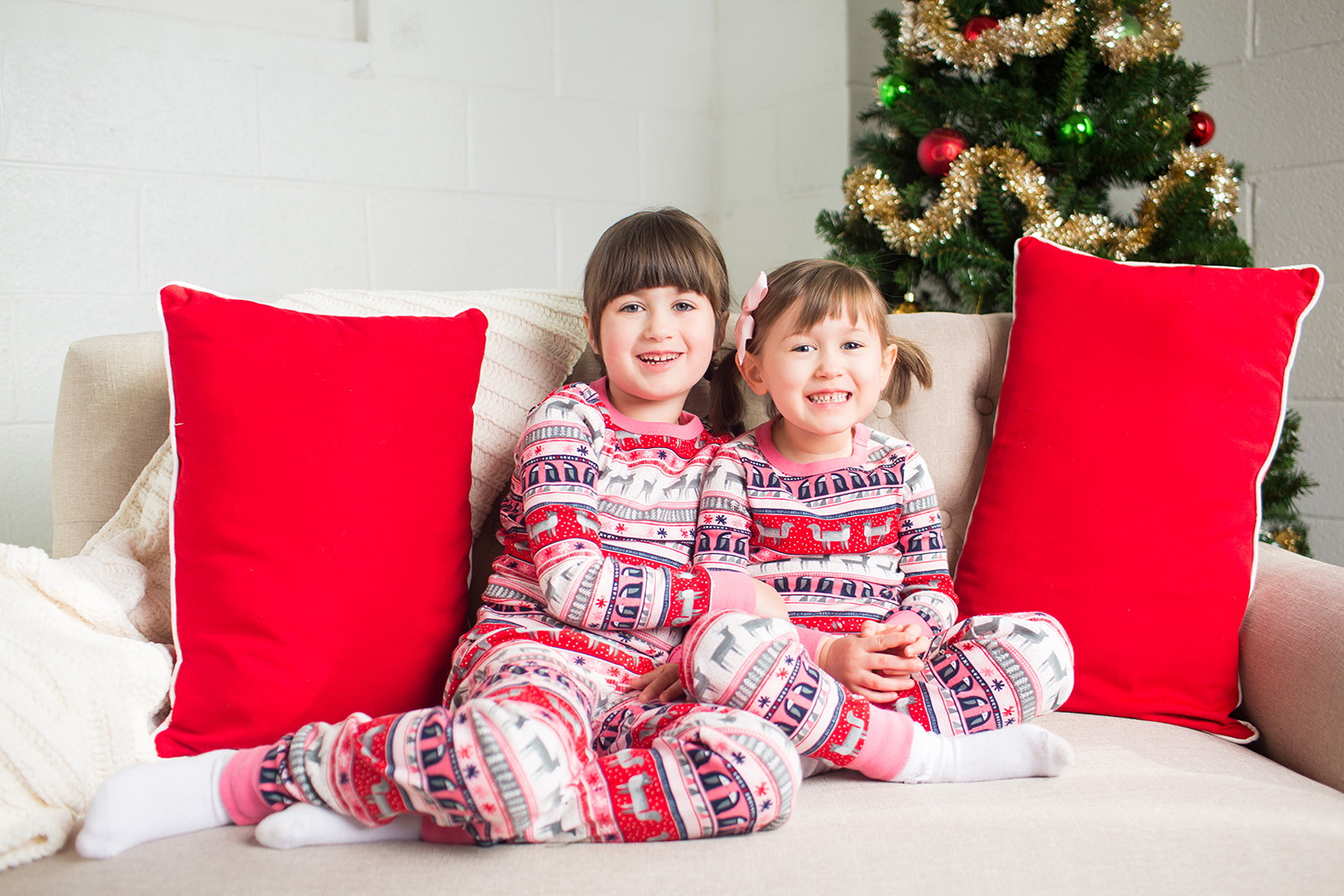 two little girls sitting on a cream couch in front of a Christmas Tree.