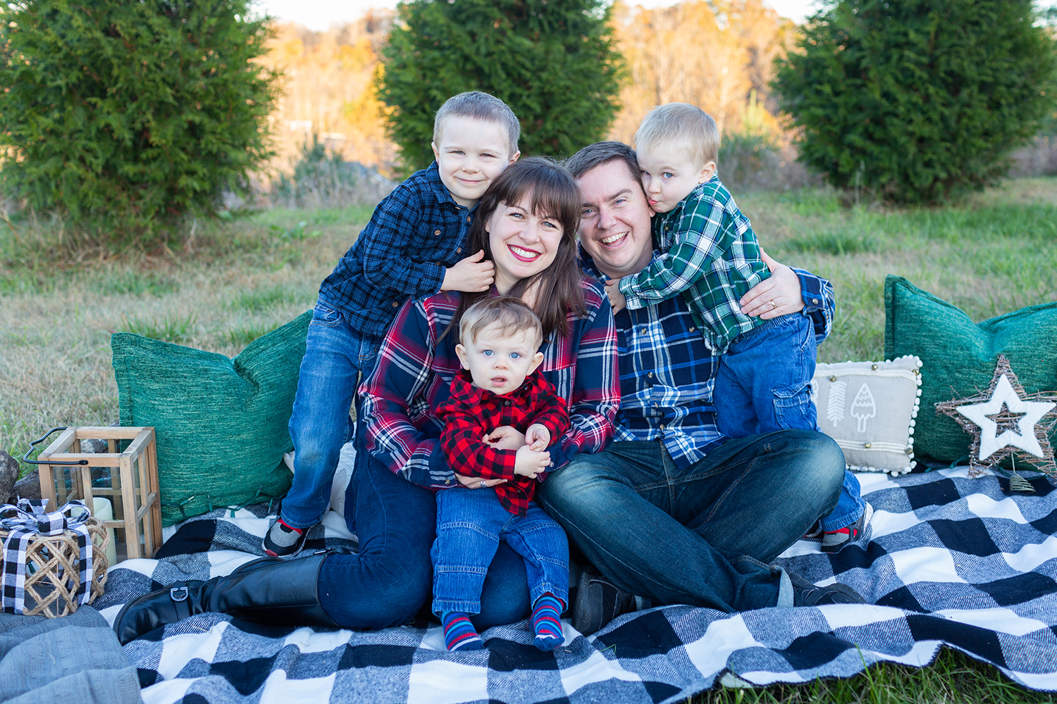 family seated on a black and white plaid blanket in a field of Christmas Trees.